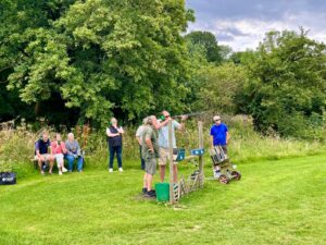 A group of people clay target shooting. One man shooting with an instructor and a small group watching him. 