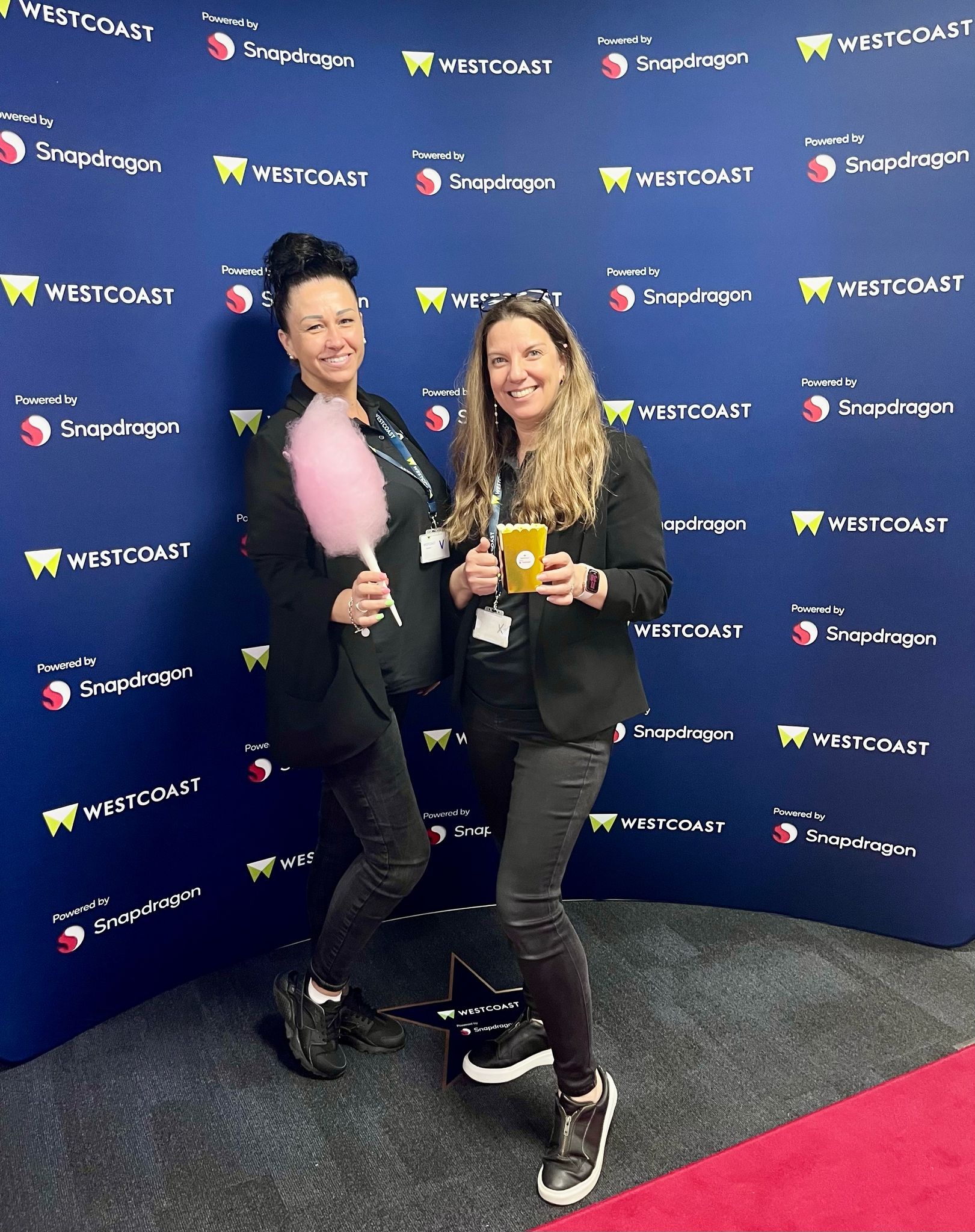 Two team members in front of a step and repeat banner. One is holding candyfloss and the other popcorn