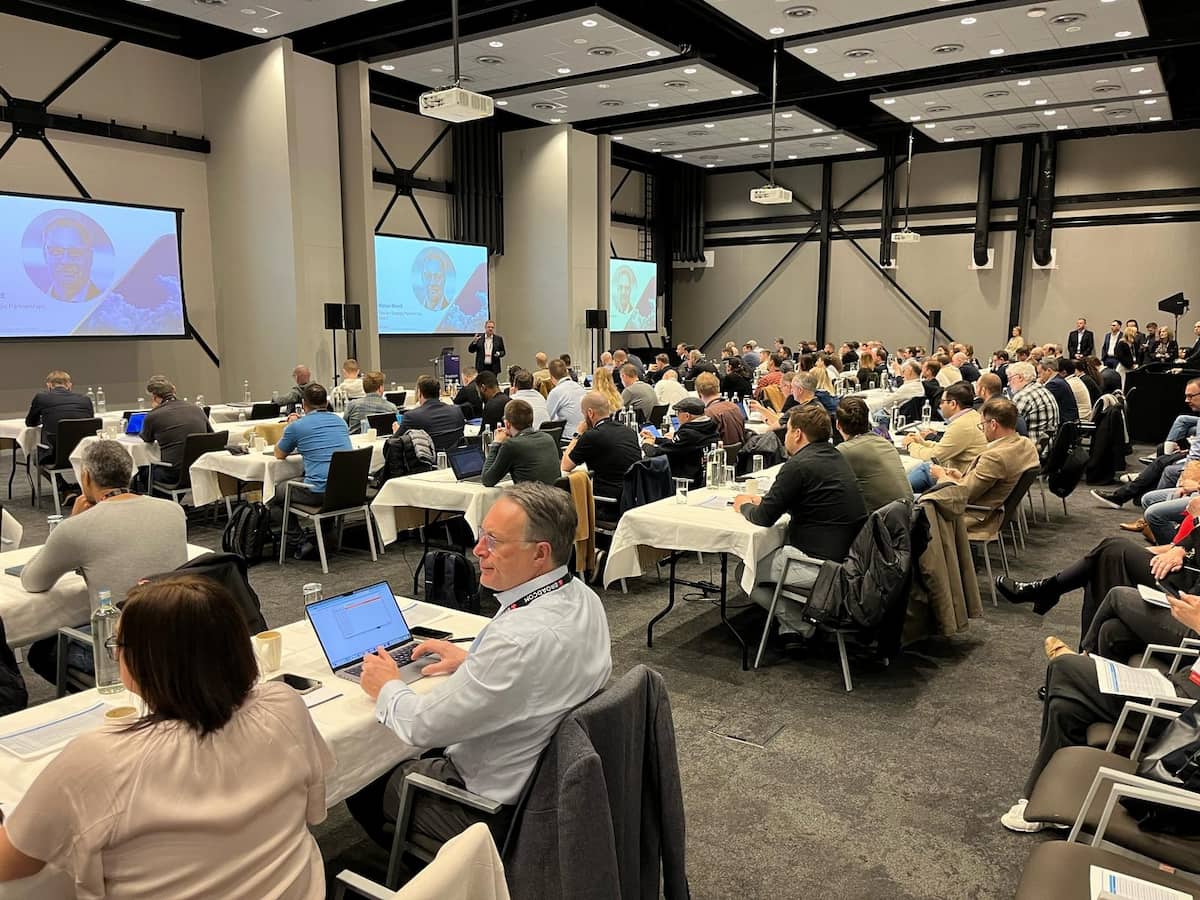 A large group of people at an event, sat on rows of tables. A speaker is stood at the front in front of a screen.